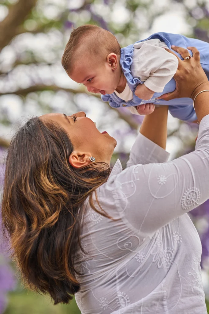 Mother holding her smiling 6-month-old baby girl under blooming jacaranda trees at Parramatta Park