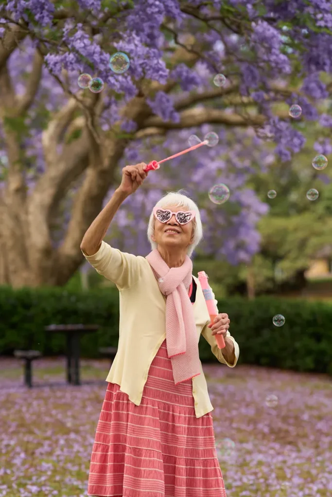 Elderly woman playing with bubbles outdoors under jacaranda